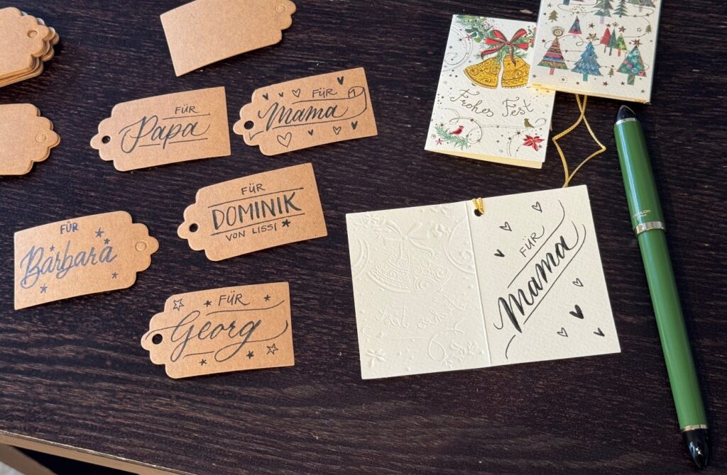 Geschenkanhänger mit handschriftlichen Namen auf einem Tisch, daneben eine grüne Füllfeder und Weihnachtskarten.